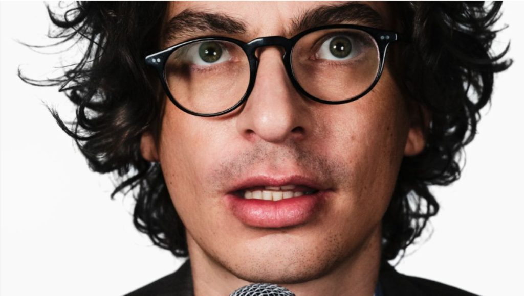 Adam Friedland seated at a retro talk-show desk with a city-light backdrop