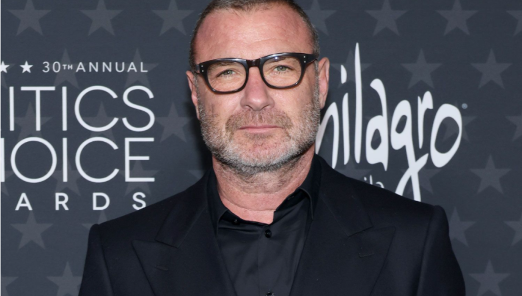 Liev Schreiber on a film set at night, city lights behind him