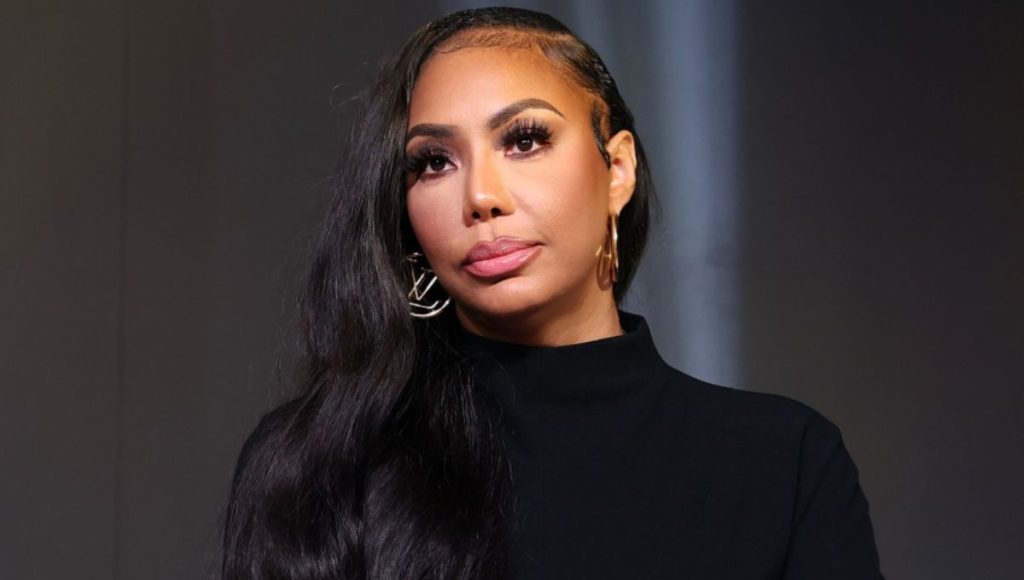 Tamar Braxton on stage in a spotlight, hand on mic, with a serious expression