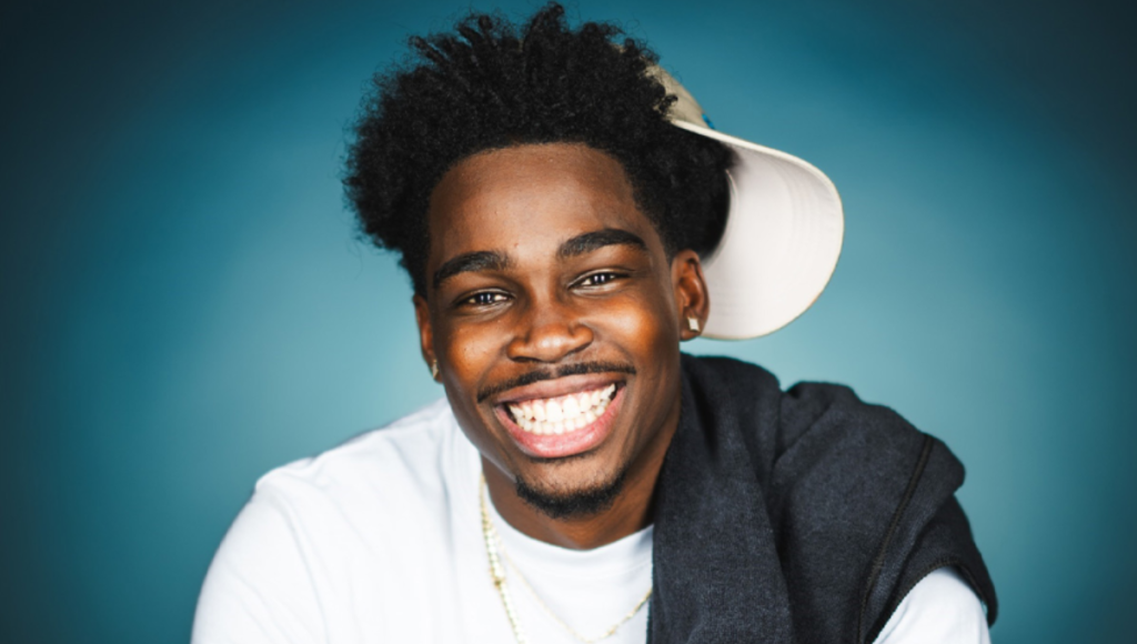 Kam Patterson performing stand-up on stage, holding a microphone, mid-joke