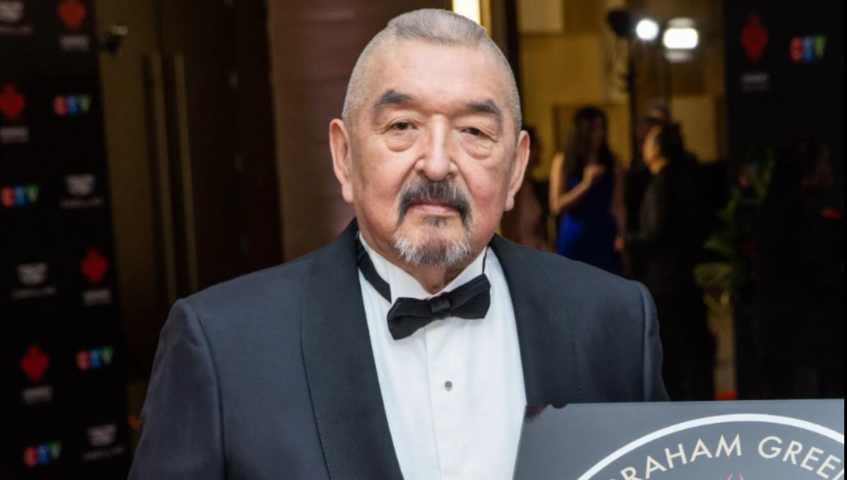 Graham Greene smiling on a red carpet in a dark suit, greeting photographers